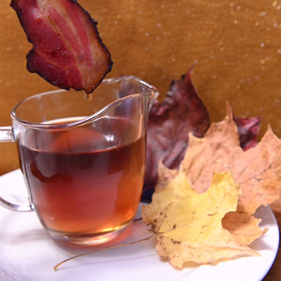 Pastured Pork Maple Bacon being dipped into a glass of syrup with autumn leaves on a white plate.
