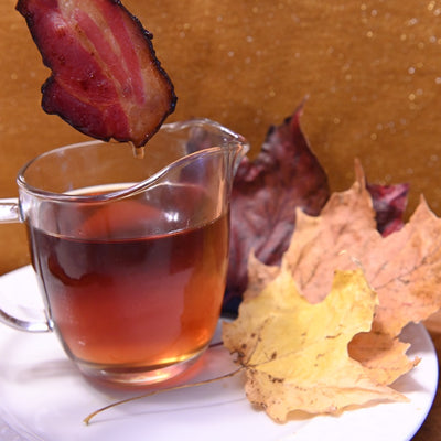 Pastured Pork Maple Bacon being dipped into a glass of syrup with autumn leaves on a white plate.